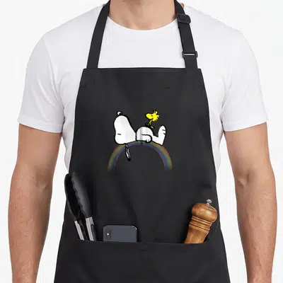 Zip rainbow apron - Snoopy lying on rainbow with yellow Woodstock on stomach, cartoon style — shown on a black cotton canvas apron against a white background
