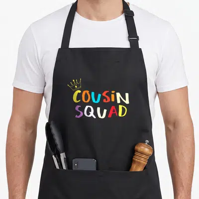 Squad crew apron - COUSIN SQUAD text in yellow orange blue red purple white with crown — shown on a black cotton canvas apron against a white background