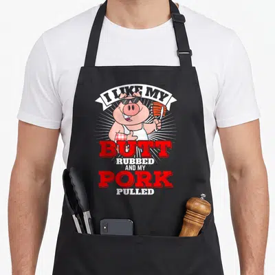 Butt pulled lover apron - Cartoon pig in sunglasses holding ribs with red and white text about pulled pork — shown on a black cotton canvas apron against a white background