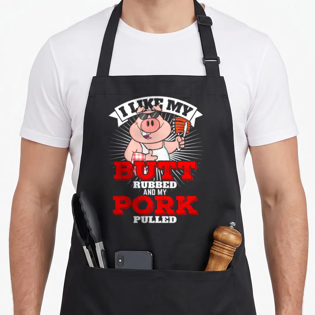 Butt pulled lover apron - Cartoon pig in sunglasses holding ribs with red and white text about pulled pork — shown on a black cotton canvas apron against a white background