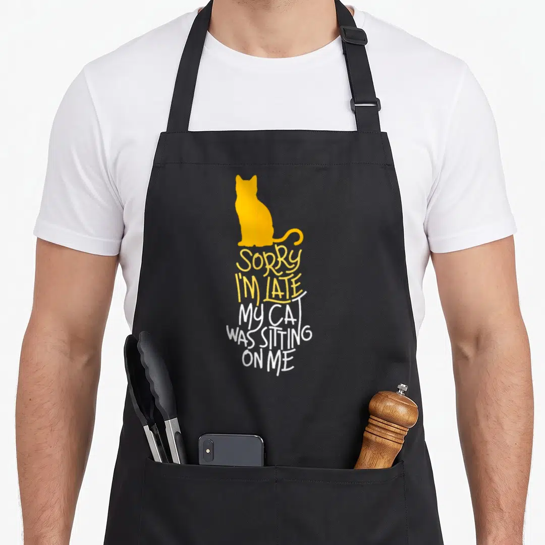 Late cat apron - Yellow cat silhouette above Sorry I'm Late My Cat Was Sitting On Me text in yellow and white — shown on a black cotton canvas apron against a white background