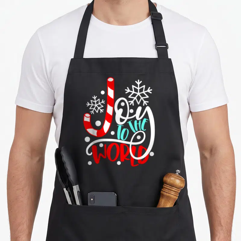 Joy candy cane apron - Candy cane shaped J in Joy TO THE WORLD text with snowflakes and light blue accents — shown on a black cotton canvas apron against a white background