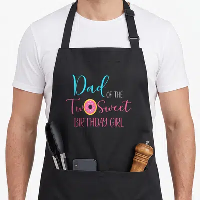 Cute sweet birthday apron - Dad OF THE Two Sweet BIRTHDAY GIRL text with pink donut replacing the O in Two — shown on a black cotton canvas apron against a white background