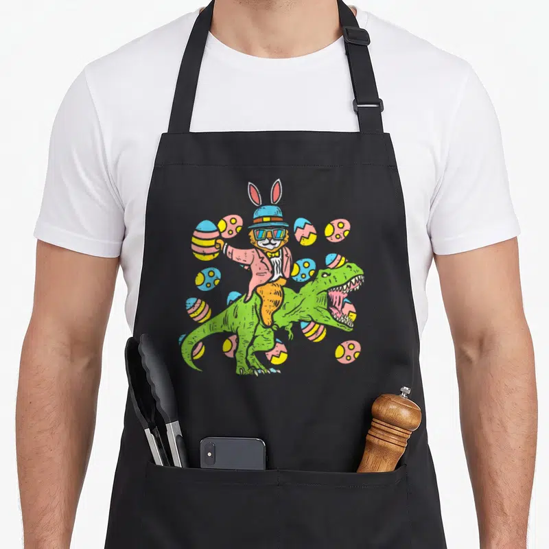 Cat easter bunny aprons - Cartoon rabbit in pink suit riding green dinosaur among blue yellow and orange eggs — shown on a black cotton canvas apron against a white background