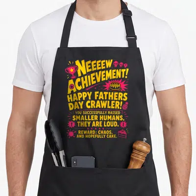 Crawler father day apron - Yellow and pink text reading Neeeew Achievement with trophy, skulls, and cake graphics — shown on a black cotton canvas apron against a white background