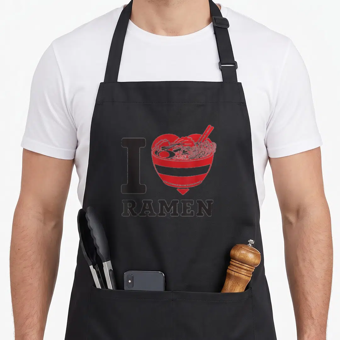 Noodle i love ramen apron - Red heart-shaped ramen bowl with chopsticks and egg next to black I and RAMEN text — shown on a black cotton canvas apron against a white background