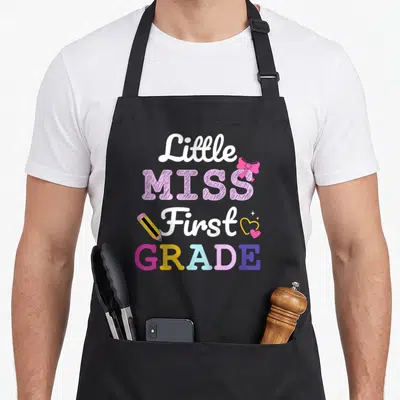 1st school miss grade apron - Little MISS First GRADE text with pink bow, pencil, and hearts in white pink yellow purple — shown on a black cotton canvas apron against a white background