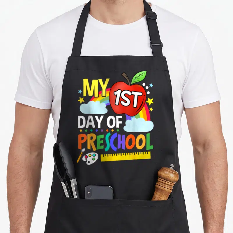 Colorful preschool apron - Red apple with 1ST text, rainbow, clouds, and art supplies below MY 1ST DAY OF PRESCHOOL — shown on a black cotton canvas apron against a white background