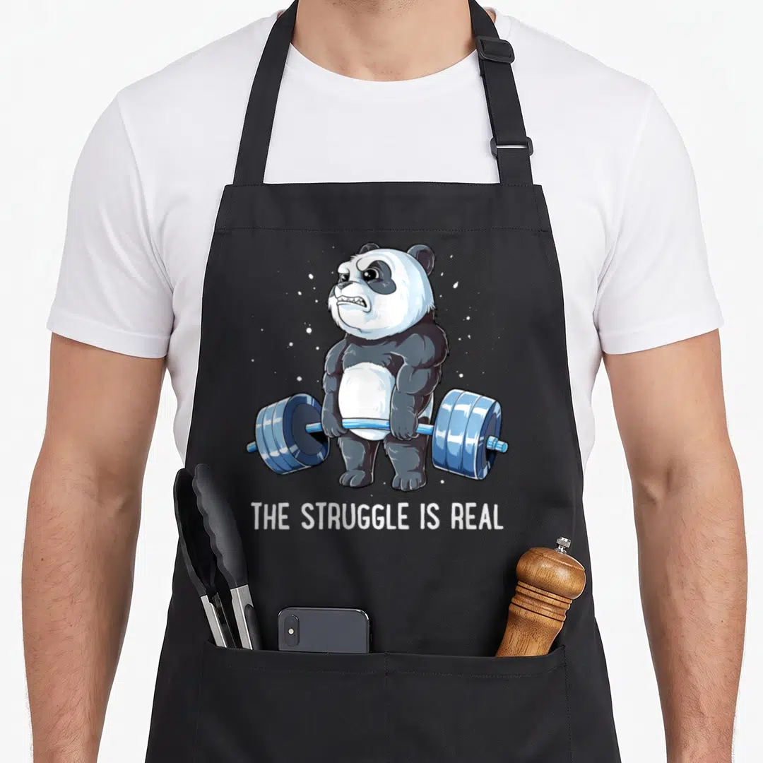 Panda deadlift apron - Muscular cartoon panda lifting blue barbell with text THE STRUGGLE IS REAL — shown on a black cotton canvas apron against a white background
