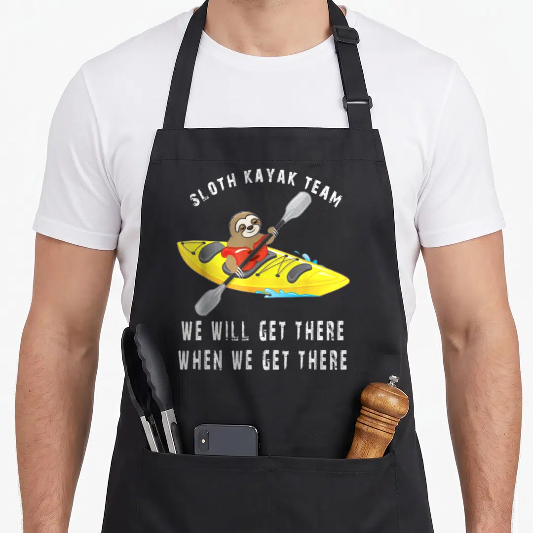 Paddling sloth apron - Cartoon sloth in yellow kayak wearing red shirt with SLOTH KAYAK TEAM text — shown on a black cotton canvas apron against a white background