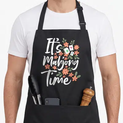 Gambling mahjong apron - It's Mahjong Time text with peach flowers and three distinct tiles in cartoon style — shown on a black cotton canvas apron against a white background