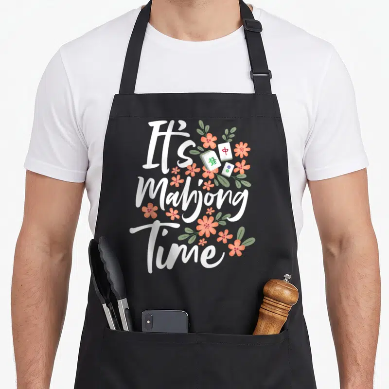 Gambling mahjong apron - It's Mahjong Time text with peach flowers and three distinct tiles in cartoon style — shown on a black cotton canvas apron against a white background