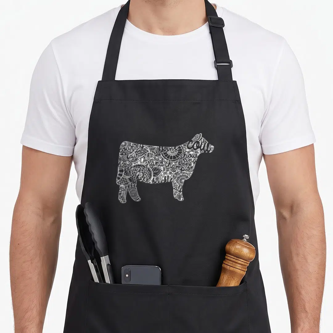 Cow stock aprons - White cow silhouette filled with doodle text including grand champion, show ring, and ribbons — shown on a black cotton canvas apron against a white background