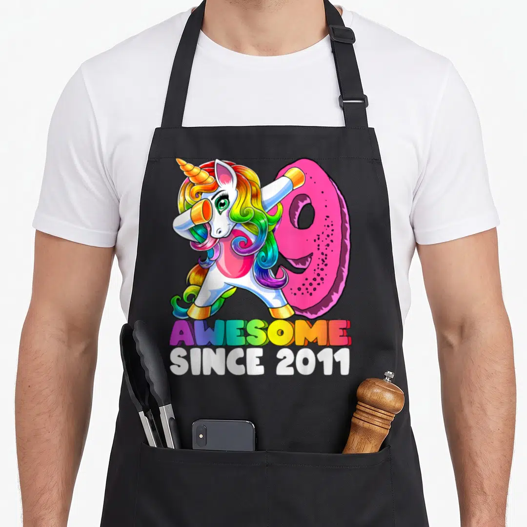 Unicorn 9th birthday apron - Dabbing unicorn in front of pink 9 with rainbow AWESOME SINCE 2011 text — shown on a black cotton canvas apron against a white background