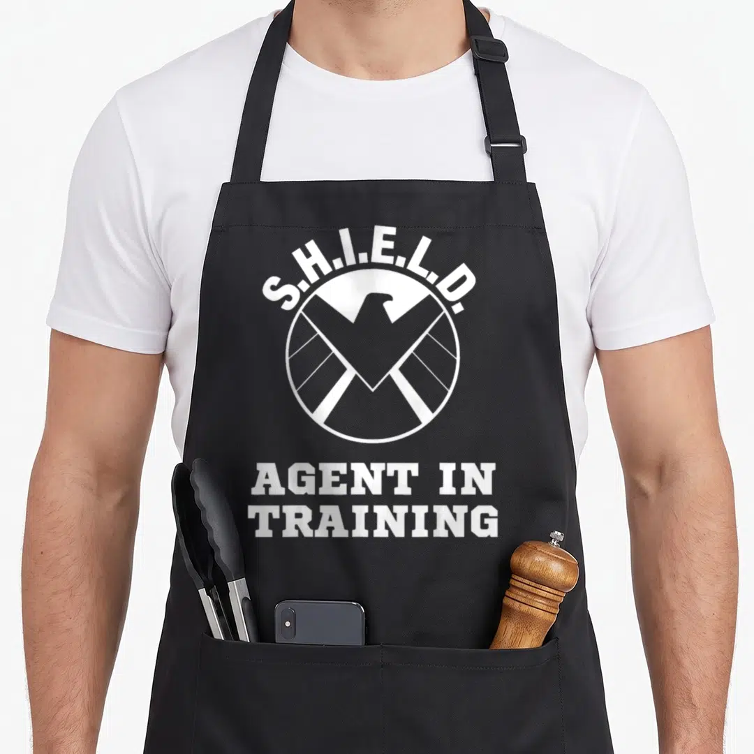 Training eagles apron - White S.H.I.E.L.D. eagle logo and Agent In Training text on black fabric — shown on a black cotton canvas apron against a white background