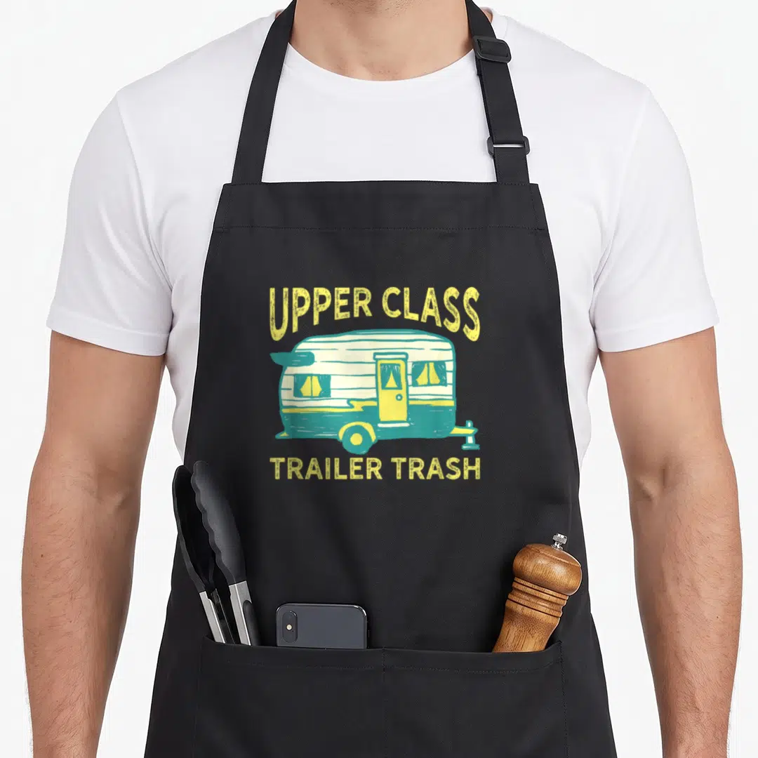 Trash trailer apron - Cartoon trailer between UPPER CLASS and TRAILER TRASH text in yellow teal white — shown on a black cotton canvas apron against a white background