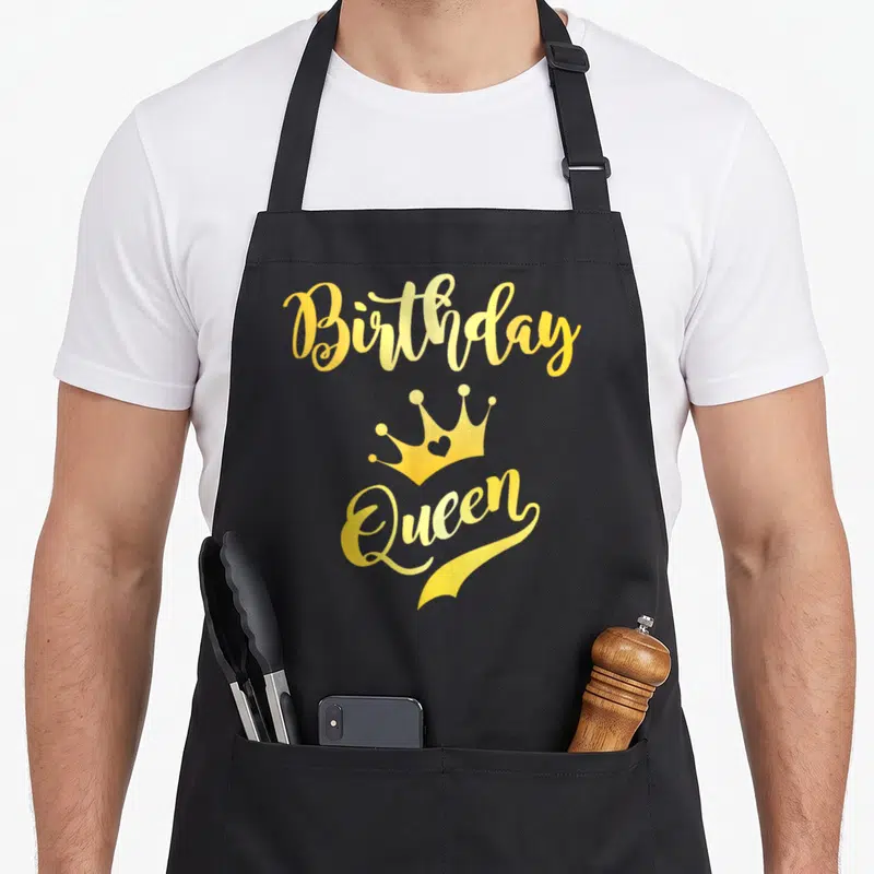 Vintage birthday queen of baking apron - Gold Birthday Queen text with a crown containing a heart — shown on a black cotton canvas apron against a white background