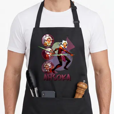 Destiny force life apron - Ahsoka Tano cartoon with dual green sabers and three circular face portraits above maroon — shown on a black cotton canvas apron against a white background