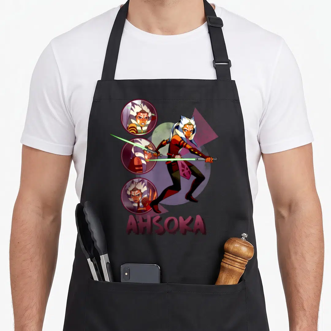 Destiny force life apron - Ahsoka Tano cartoon with dual green sabers and three circular face portraits above maroon — shown on a black cotton canvas apron against a white background
