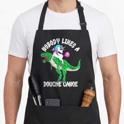 Nobody dinosaur apron - Cartoon green dinosaur with unicorn rider and white text nobody likes a douche canoe — shown on a black cotton canvas apron against a white background