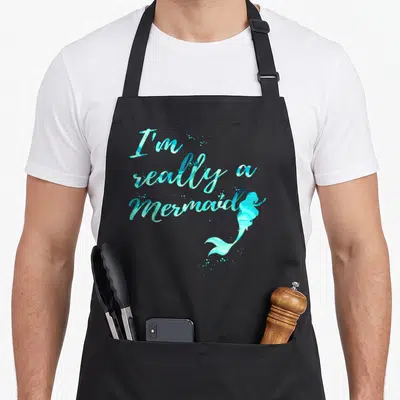 Cute mermaid apron - I'm really a Mermaid script with teal watercolor tail curving upward from bottom right — shown on a black cotton canvas apron against a white background