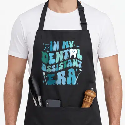 Era dentist apron - Bubbly blue text reading IN MY DENTAL ASSISTANT ERA with toothbrush, mirror, probe and smiling — shown on a black cotton canvas apron against a white background