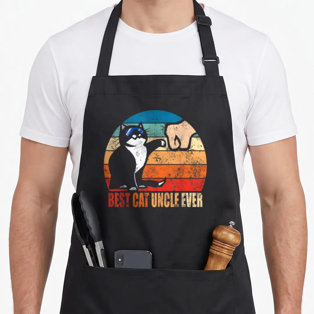 Uncle cat apron - Retro cat silhouette fist bumping human hand against striped sunset circle with BEST CAT UNCLE EVER — shown on a black cotton canvas apron against a white background
