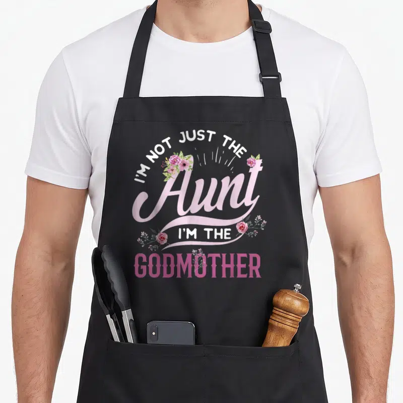 Aunt godmother apron - Pink and white text reading I'm Not Just The Aunt I'm The Godmother surrounded by green and pink — shown on a black cotton canvas apron against a white background