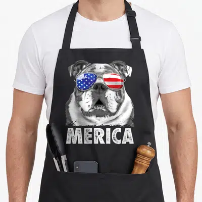 4th english chef aprons - Realistic bulldog head with USA flag aviator sunglasses and distressed MERICA text below — shown on a black cotton canvas apron against a white background