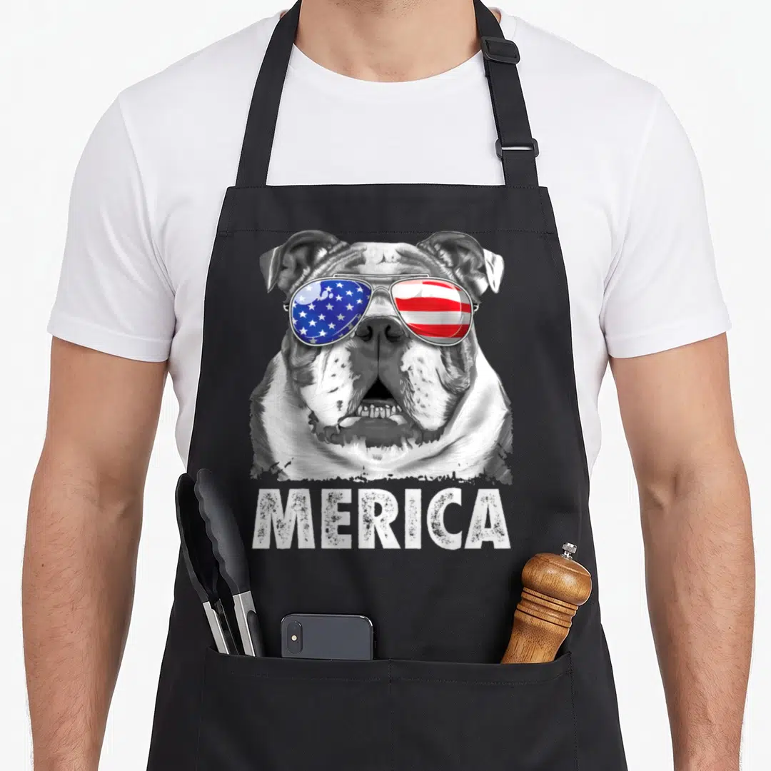 4th english chef aprons - Realistic bulldog head with USA flag aviator sunglasses and distressed MERICA text below — shown on a black cotton canvas apron against a white background