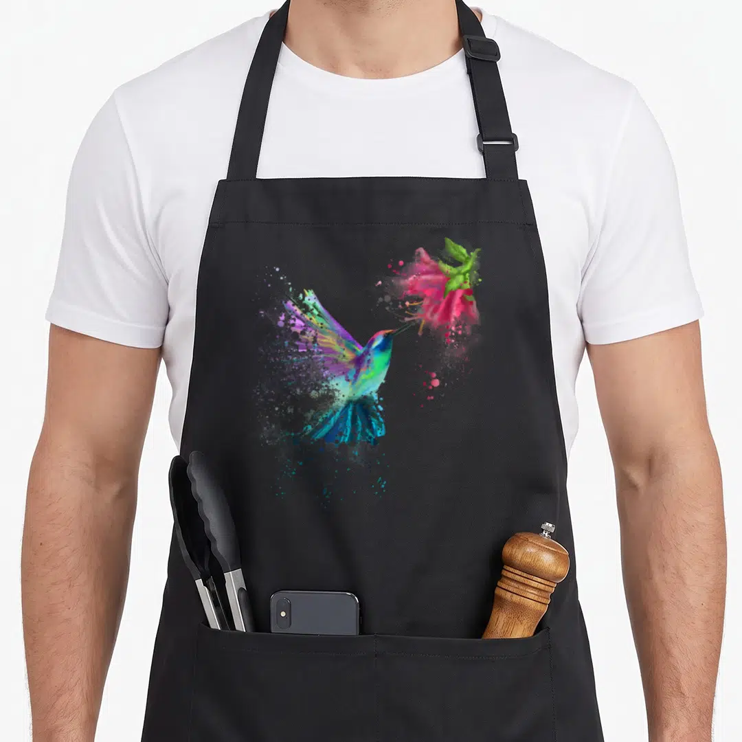 Flower bird apron - Hummingbird flying to pink flower with blue green purple pink watercolor splatters — shown on a black cotton canvas apron against a white background