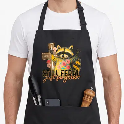 Raccoon christian apron - Cartoon raccoon with pink flowers standing in front of a wooden cross and text reading Still — shown on a black cotton canvas apron against a white background