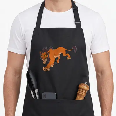 Prowling lion apron - Crouching cartoon lion in orange and brown with green eyes on black background — shown on a black cotton canvas apron against a white background