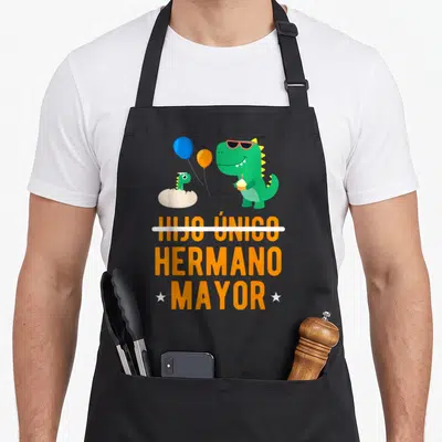 Hijo mayor apron - Green dinosaur with sunglasses holding cupcake above orange text HIJO ÚNICO crossed out HERMANO — shown on a black cotton canvas apron against a white background