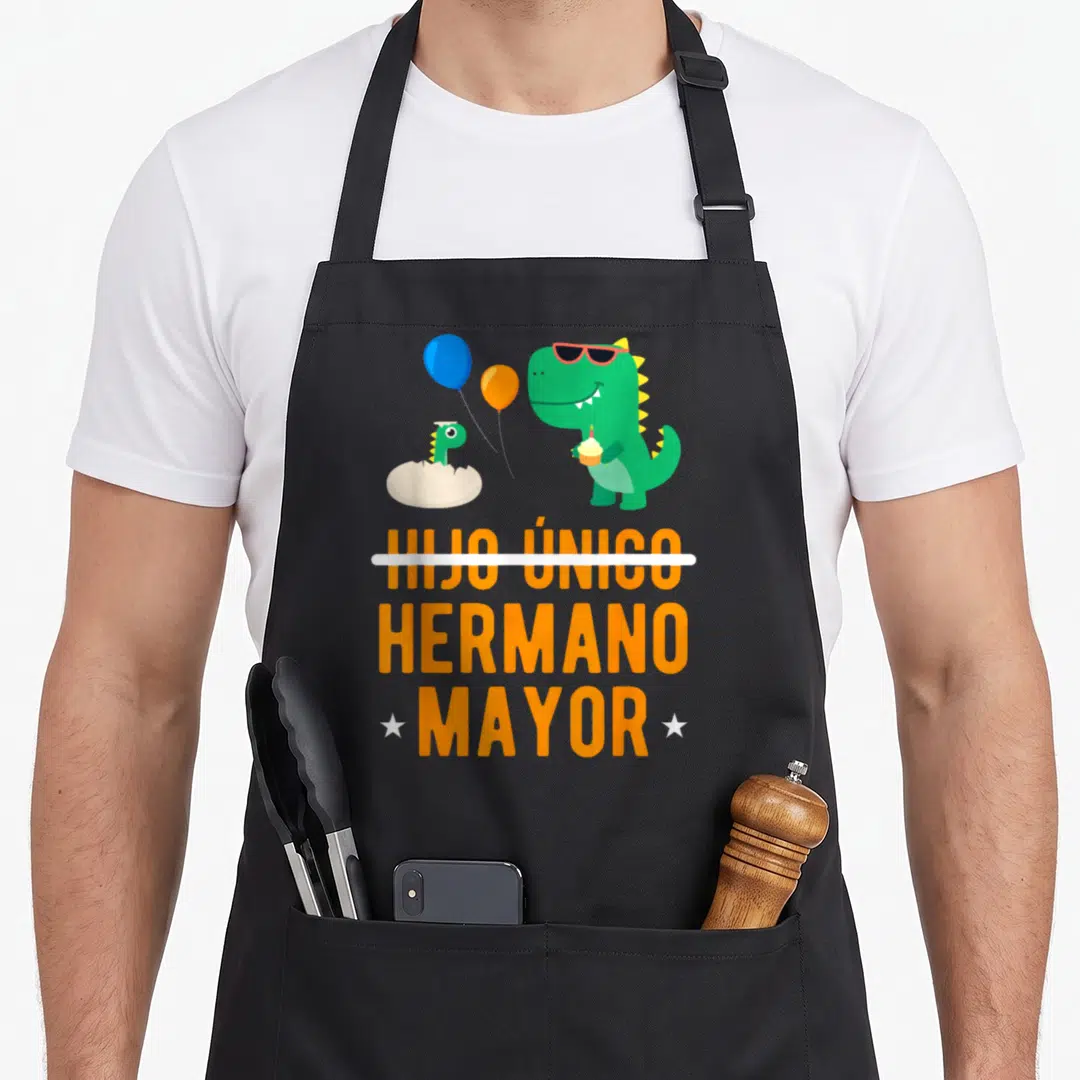 Hijo mayor apron - Green dinosaur with sunglasses holding cupcake above orange text HIJO ÚNICO crossed out HERMANO — shown on a black cotton canvas apron against a white background