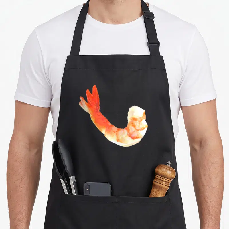 Shrimp cocktail apron - Single photo-style shrimp in orange and white centered on fabric — shown on a black cotton canvas apron against a white background