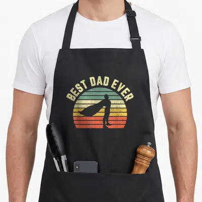 Superhero father day apron - Retro sun with BEST DAD EVER text and grey cape silhouette — shown on a black cotton canvas apron against a white background