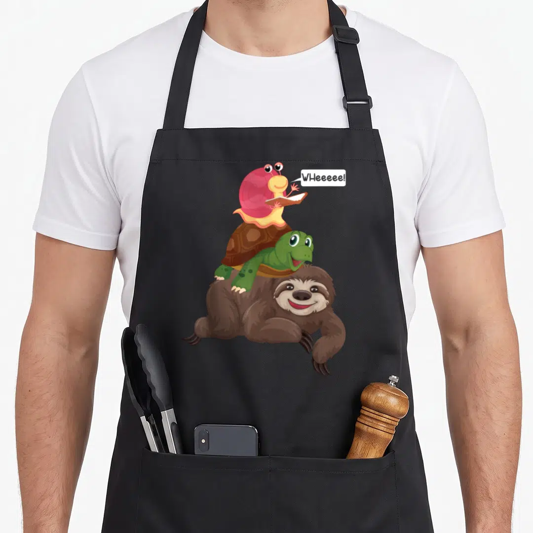 Piggyback sloth apron - Cartoon pink snail with book on green turtle atop brown sloth with WHeeeee speech bubble — shown on a black cotton canvas apron against a white background
