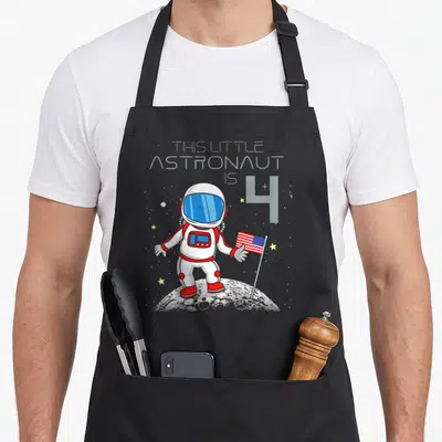 Geek birthday apron - Cartoon astronaut holding USA flag on grey moon with text THIS LITTLE ASTRONAUT IS 4 — shown on a black cotton canvas apron against a white background