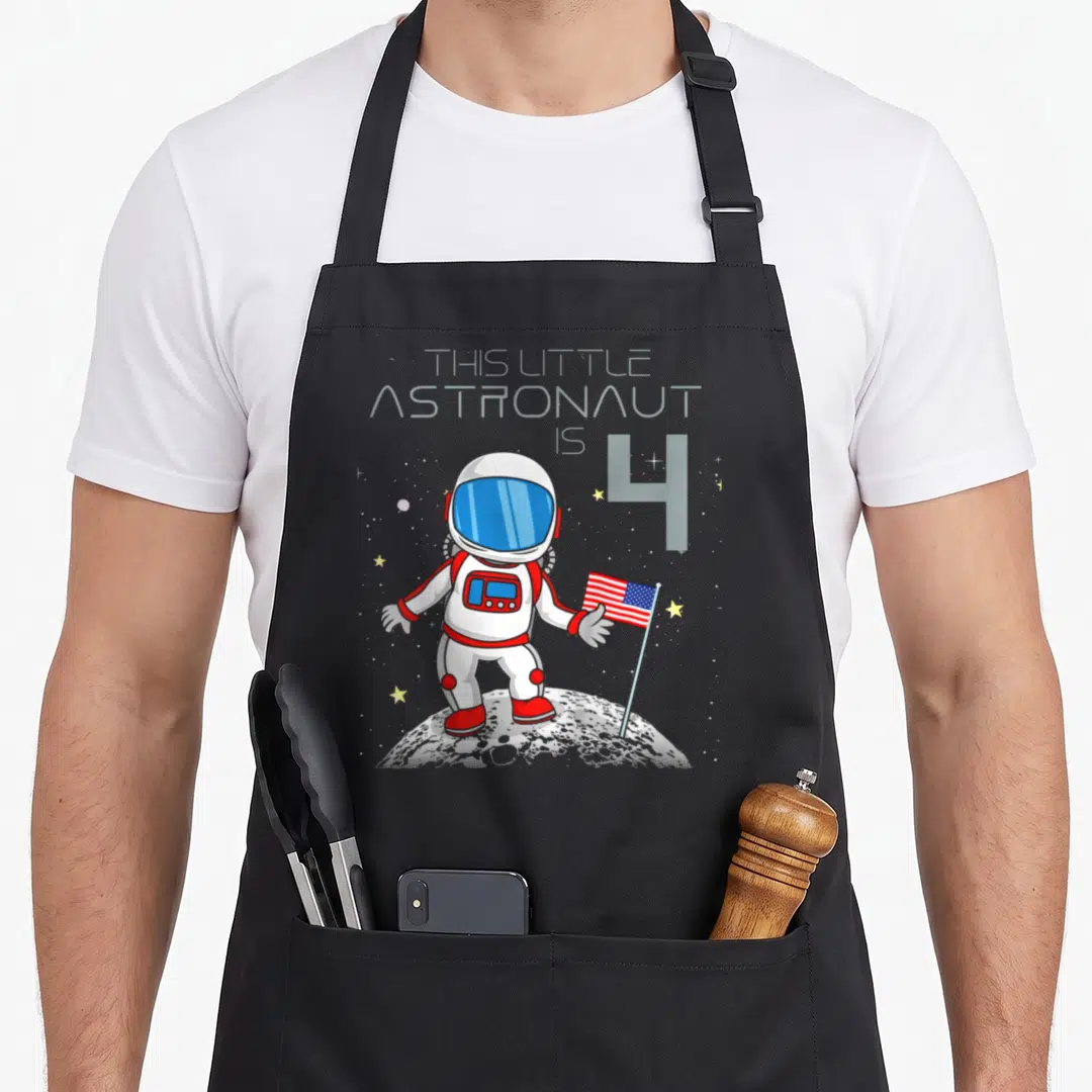 Geek birthday apron - Cartoon astronaut holding USA flag on grey moon with text THIS LITTLE ASTRONAUT IS 4 — shown on a black cotton canvas apron against a white background