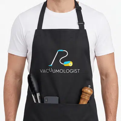 Neat cleaner apron - Cartoon vacuum with blue and yellow details above white Vacuumologist text on black background — shown on a black cotton canvas apron against a white background