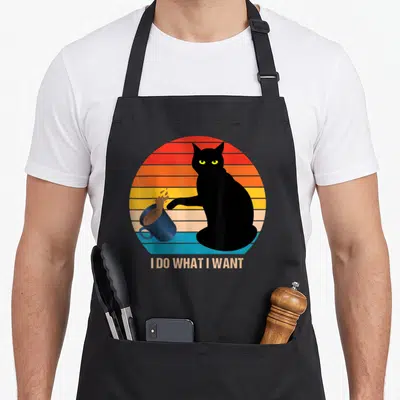 Funny cat apron for hosting - Black cat silhouette knocking over blue mug in front of striped retro sun with text below — shown on a black cotton canvas apron against a white background