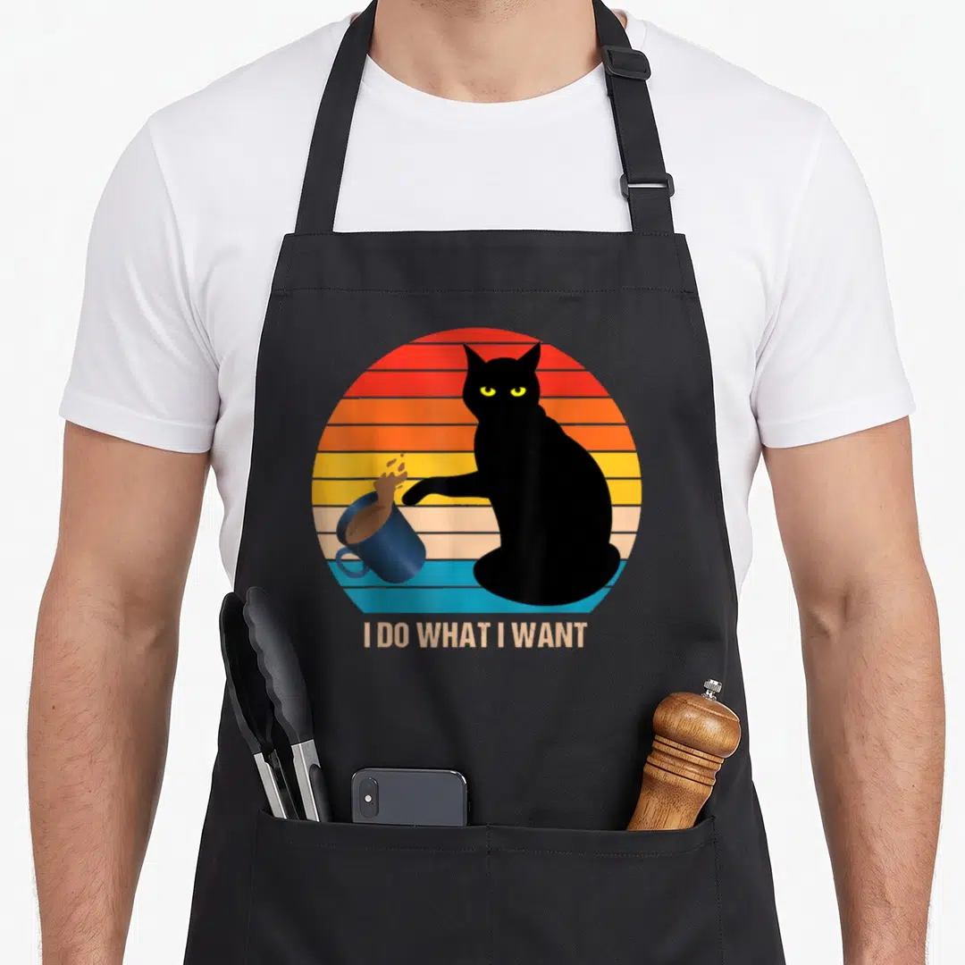 Funny cat apron for hosting - Black cat silhouette knocking over blue mug in front of striped retro sun with text below — shown on a black cotton canvas apron against a white background