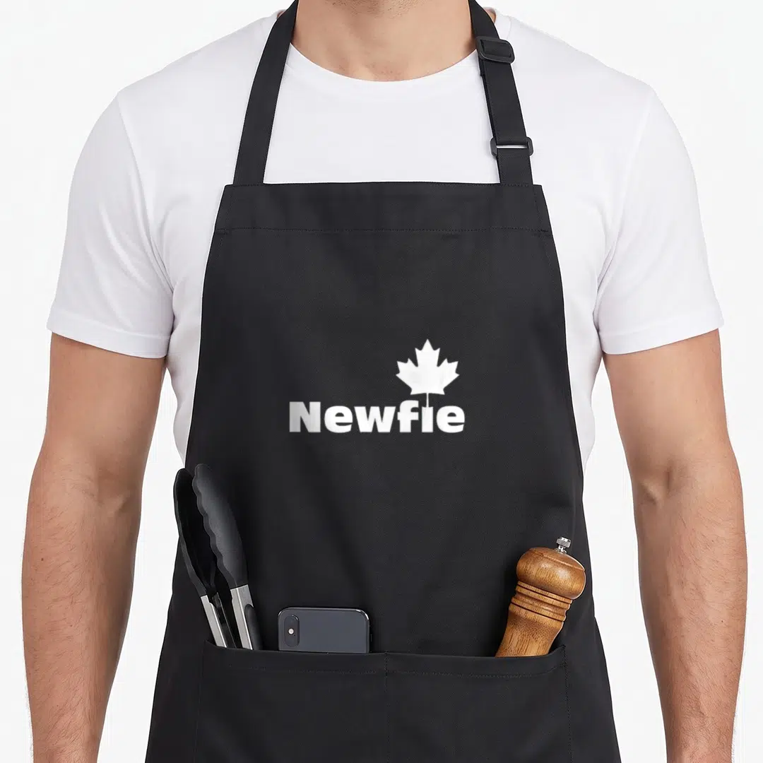 Newfie pride apron - White maple leaf centered above Newfie text on dark background — shown on a black cotton canvas apron against a white background