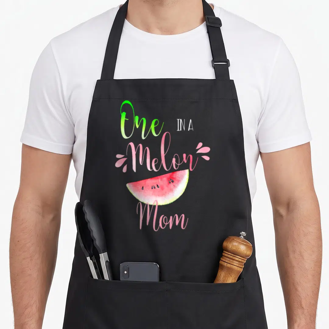 Cute melon birthday apron - Watercolor watermelon slice with One IN A Melon Mom text in pink and green — shown on a black cotton canvas apron against a white background
