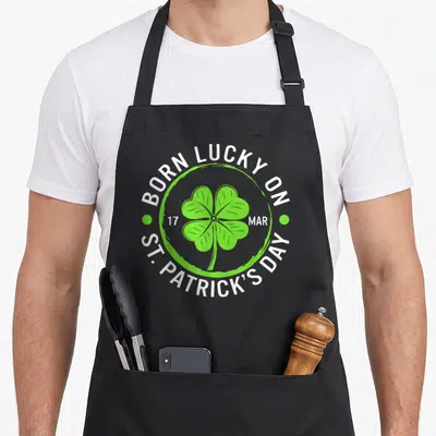 March irish st patrick day aprons - Green four leaf clover inside circle with Born Lucky On St Patricks Day text — shown on a black cotton canvas apron against a white background