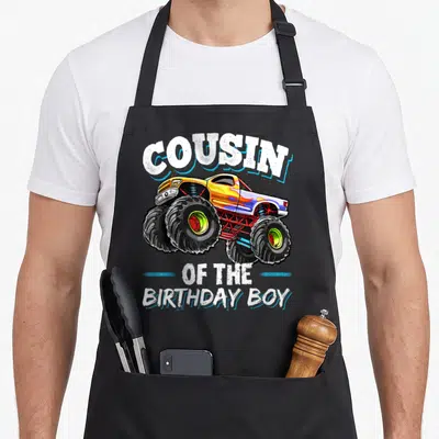 Cousin truck apron - Monster truck illustration with cousin of the birthday boy text in yellow white black red — shown on a black cotton canvas apron against a white background
