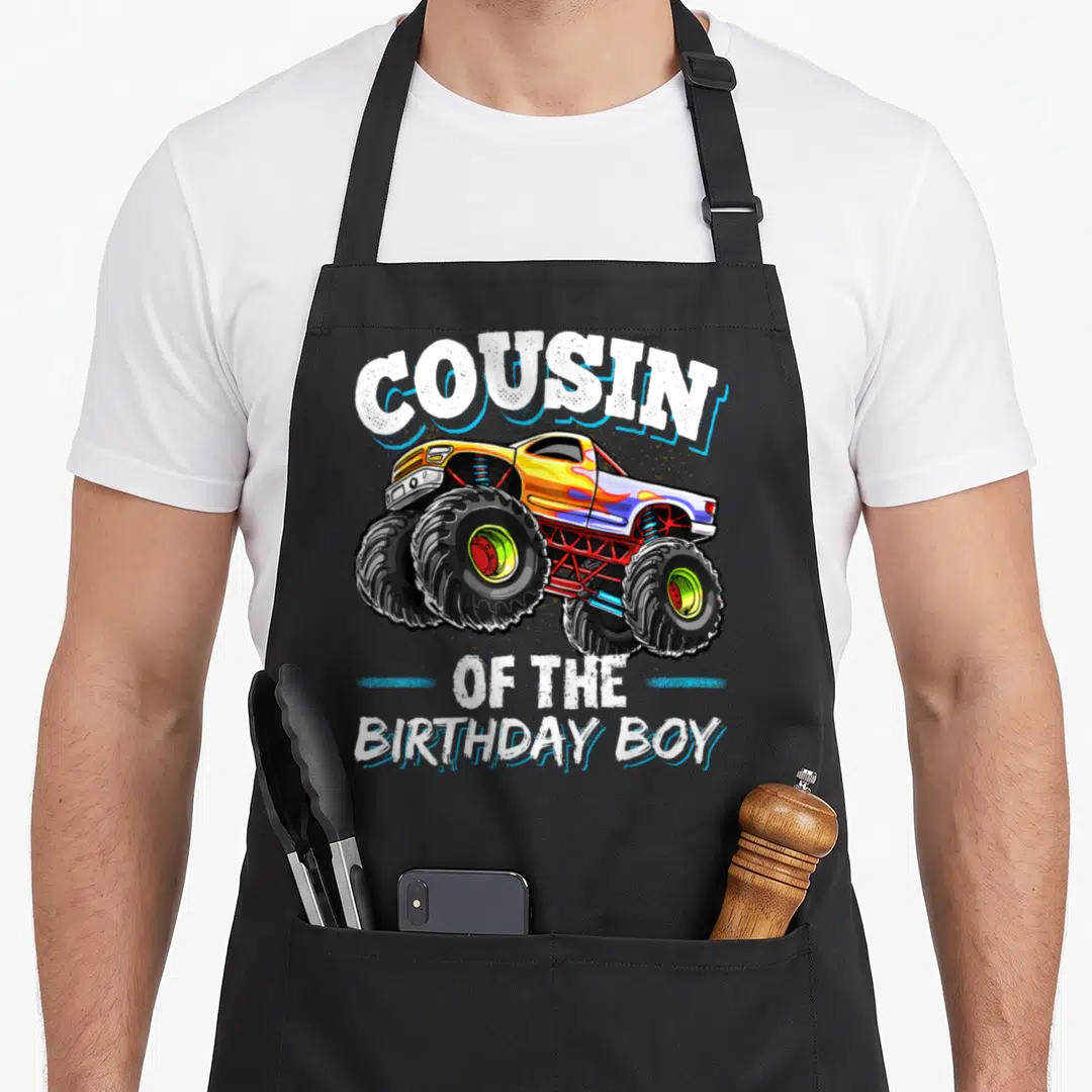 Cousin truck apron - Monster truck illustration with cousin of the birthday boy text in yellow white black red — shown on a black cotton canvas apron against a white background