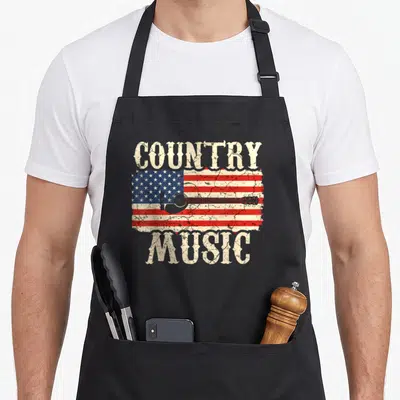 Music cook's country aprons - Distressed USA flag with guitar silhouette and beige COUNTRY MUSIC text — shown on a black cotton canvas apron against a white background