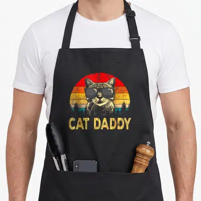 Retro daddy cat dad apron - Cat wearing sunglasses in front of red orange yellow and teal striped sun with text CAT — shown on a black cotton canvas apron against a white background